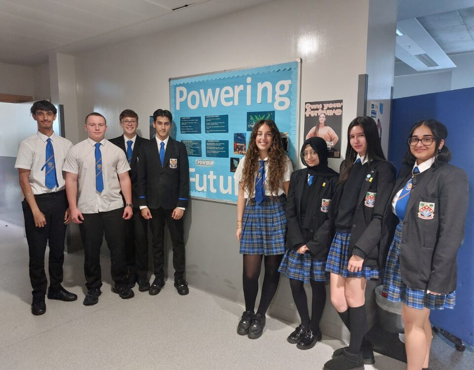 Eight high school students in uniform stand smiling in a hallway next to a blue bulletin board that reads “Powering our future.” Some wear blazers while others wear shirts and ties.