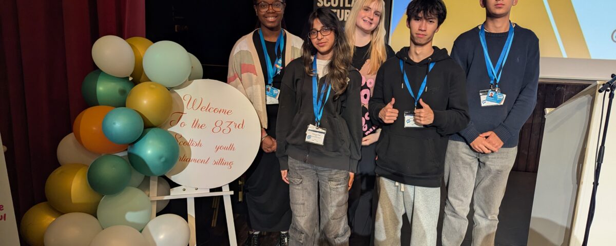 Five young people stand together on a stage next to a colorful balloon display and a sign that reads, “Welcome To the 83rd Scottish Youth Parliament Sitting.” They are smiling and wearing name badges.