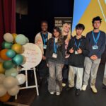 Five young people stand together on a stage next to a colorful balloon display and a sign that reads, “Welcome To the 83rd Scottish Youth Parliament Sitting.” They are smiling and wearing name badges.