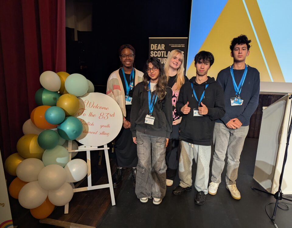 Five young people stand together on a stage next to a colorful balloon display and a sign that reads, “Welcome To the 83rd Scottish Youth Parliament Sitting.” They are smiling and wearing name badges.