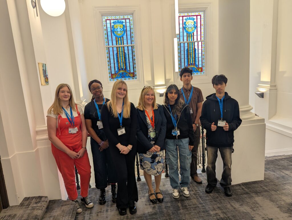 A group of seven people, including teenagers and one adult, stand indoors in front of two stained glass windows. They all wear name badges and blue lanyards, smiling for the photo.