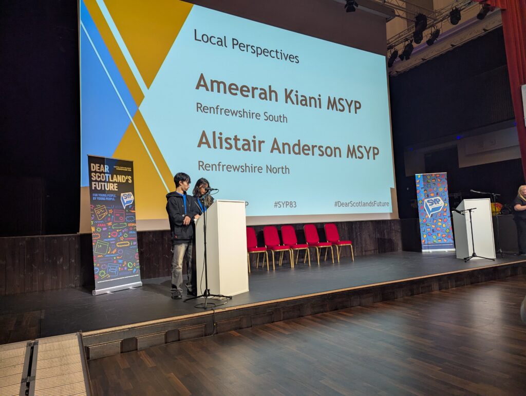 Two young people stand at a podium on stage, presenting to an audience. A large screen behind them displays the names Ameerah Kiani and Alistair Anderson as MSYP speakers for Renfrewshire.