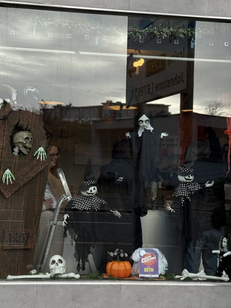 A Halloween display in a window features skeletons, ghosts, a coffin, a pumpkin, and spooky decorations. A person is partially visible inside behind the display.