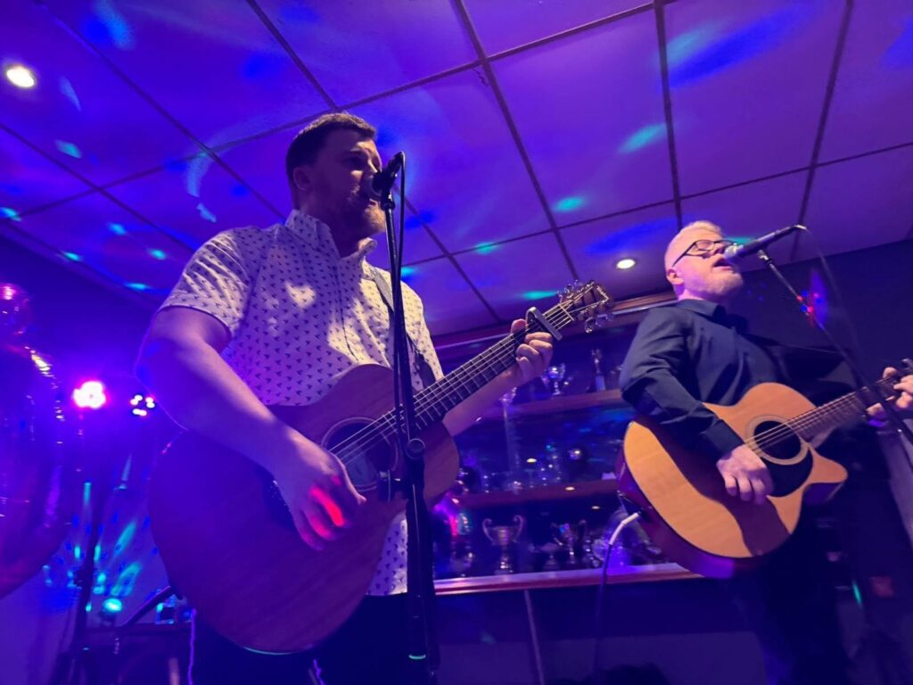 Two men play acoustic guitars and sing into microphones on stage, illuminated by colorful purple and blue lights. Trophies are displayed on shelves behind them, creating a lively and celebratory atmosphere.