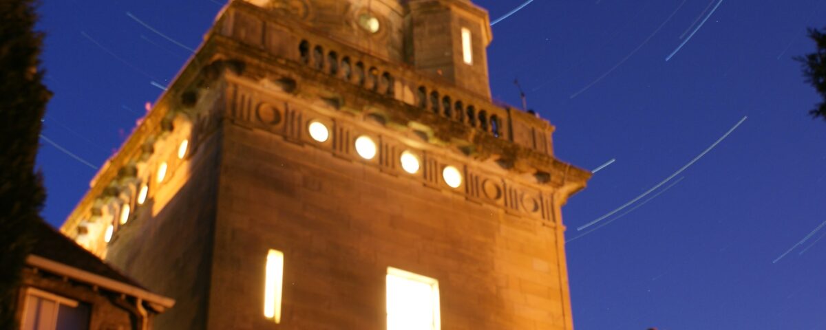 A long-exposure night photo of an observatory building, with warm yellow lights shining from its windows and circular star trails swirling in the deep blue sky above.