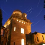 A long-exposure night photo of an observatory building, with warm yellow lights shining from its windows and circular star trails swirling in the deep blue sky above.