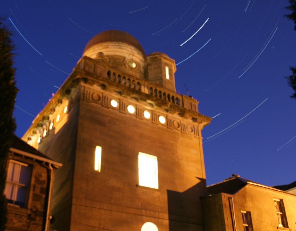 A long-exposure night photo of an observatory building, with warm yellow lights shining from its windows and circular star trails swirling in the deep blue sky above.