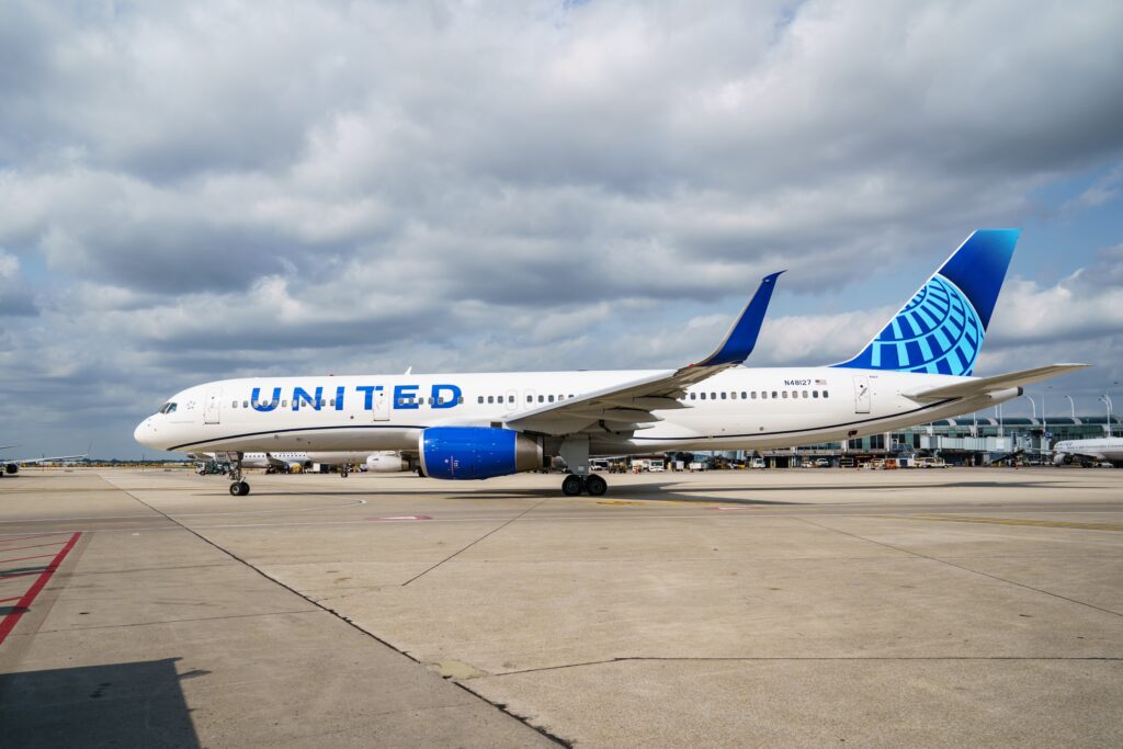 A United Airlines jet with blue and white livery is parked on the tarmac at an airport under a partly cloudy sky. Terminal buildings and jet bridges are visible in the background.