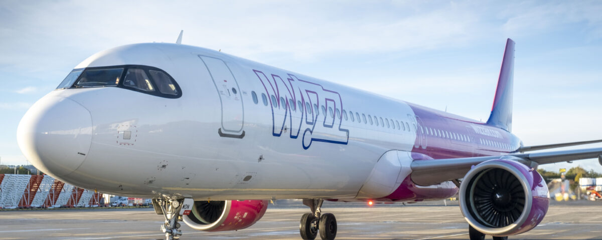 A pink and white Wizz Air passenger airplane is parked on an airport tarmac under a blue sky, with its engines and part of the terminal visible in the background.