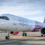 A pink and white Wizz Air passenger airplane is parked on an airport tarmac under a blue sky, with its engines and part of the terminal visible in the background.