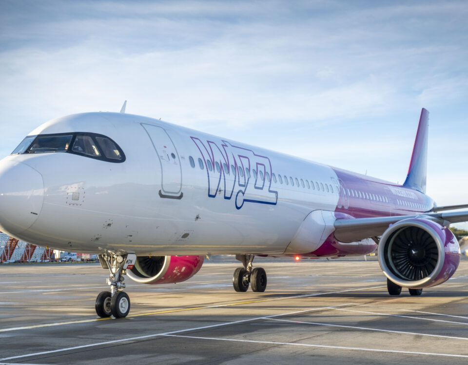 A pink and white Wizz Air passenger airplane is parked on an airport tarmac under a blue sky, with its engines and part of the terminal visible in the background.