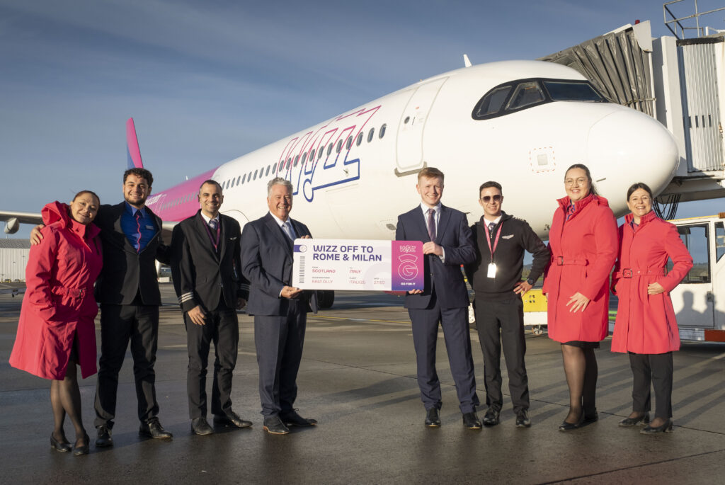 A group of eight people pose and smile in front of a Wizz Air airplane on the tarmac. Several are wearing bright red uniforms, and two people hold a large sign reading "Wizz Off to Rome & Milan.