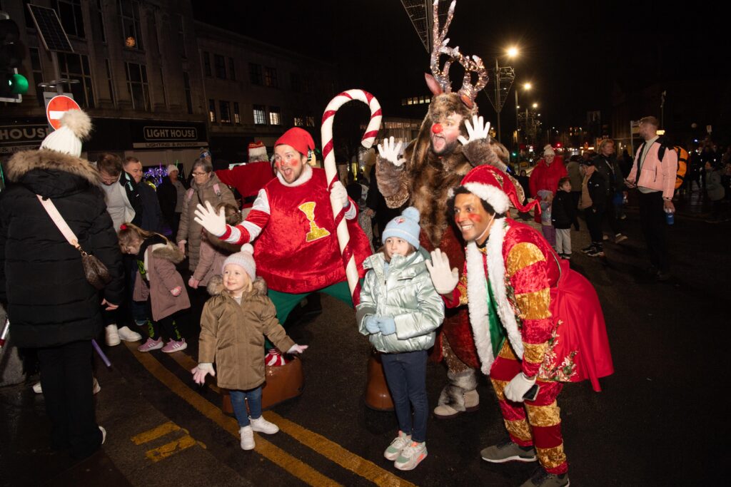 Four children pose and smile with people in festive Christmas costumes, including elves and a reindeer, at a nighttime outdoor event on a city street surrounded by other people.