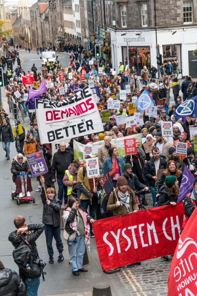 A large crowd of protesters march down a city street holding signs and banners, including ones reading “System Change” and “Demands Better.” People of various ages participate, surrounded by old stone buildings.