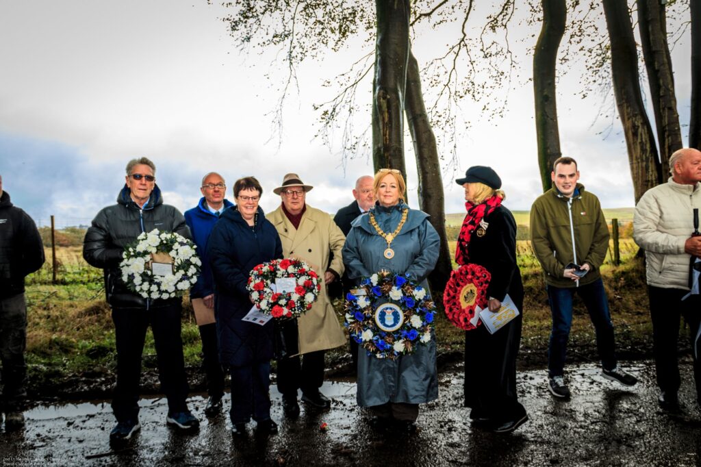 A group of people stand outdoors on a wet road, holding wreaths of white, red, and blue flowers. Tall trees and a cloudy sky are in the background. Some people wear coats, hats, and scarves.