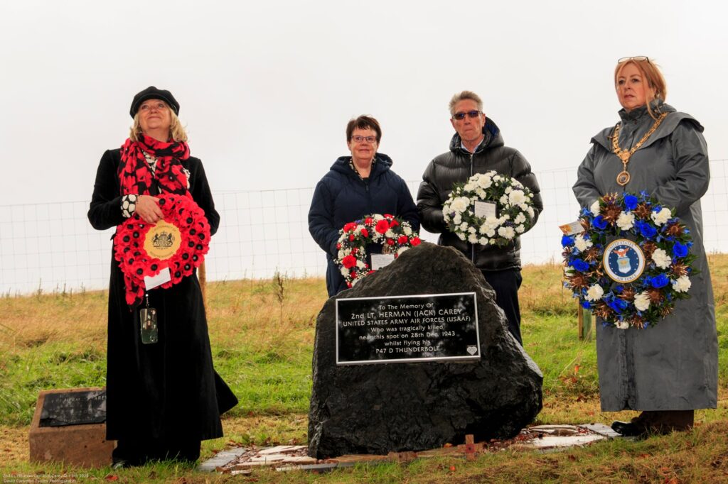 Four people stand solemnly by a memorial stone with wreaths in hand, honoring the memory of an individual. The scene is outdoors on a grassy area under an overcast sky.