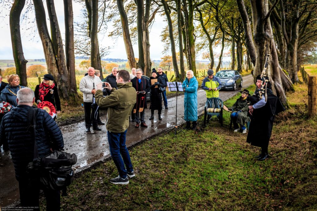 A group of people gathers on a tree-lined rural road, some holding cameras and instruments. A bagpiper in traditional dress stands to the right, while parked cars are visible in the background on a cloudy day.