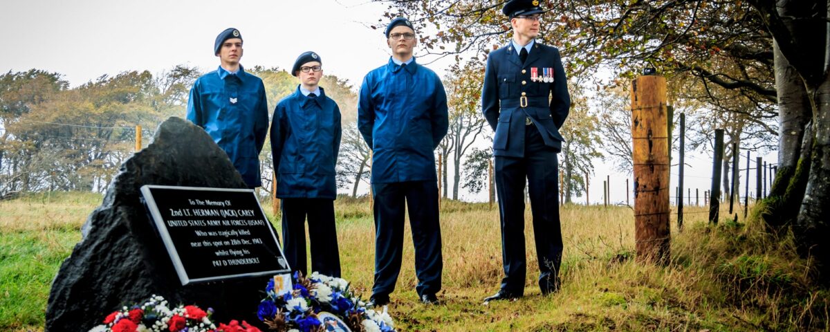 Four people stand by a memorial stone adorned with wreaths and flowers in a grassy, wooded area. Three wear blue uniforms, one wears a formal dark uniform with medals. Trees and a fence are in the background.