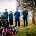 Four people stand by a memorial stone adorned with wreaths and flowers in a grassy, wooded area. Three wear blue uniforms, one wears a formal dark uniform with medals. Trees and a fence are in the background.