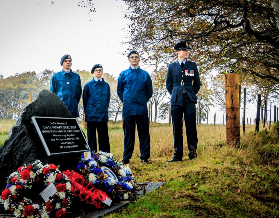 Four people stand by a memorial stone adorned with wreaths and flowers in a grassy, wooded area. Three wear blue uniforms, one wears a formal dark uniform with medals. Trees and a fence are in the background.