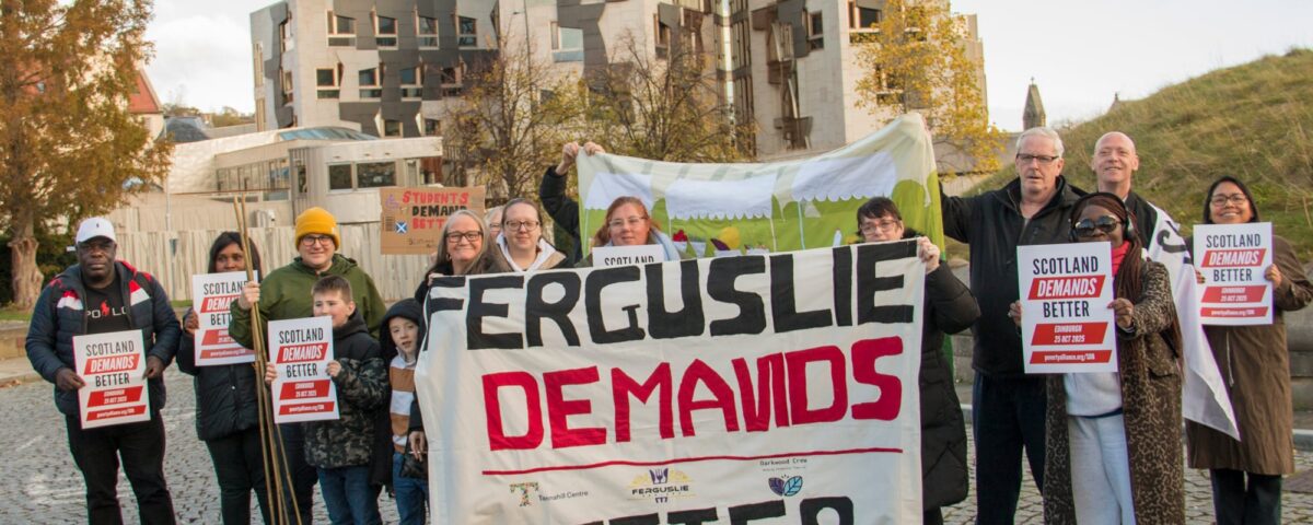 A group of people stand outside holding signs that say “Scotland Demands Better” and a large banner reading “Ferguslie Demands Better,” in front of a modern building with autumn trees nearby.