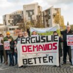 A group of people stand outside holding signs that say “Scotland Demands Better” and a large banner reading “Ferguslie Demands Better,” in front of a modern building with autumn trees nearby.