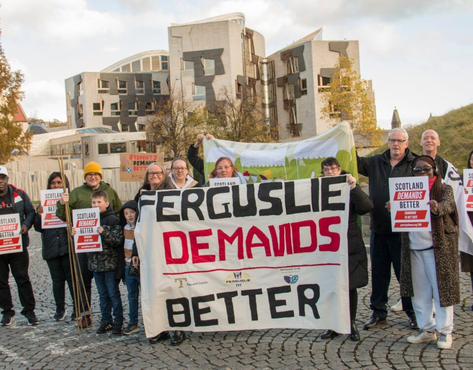 A group of people stand outside holding signs that say “Scotland Demands Better” and a large banner reading “Ferguslie Demands Better,” in front of a modern building with autumn trees nearby.