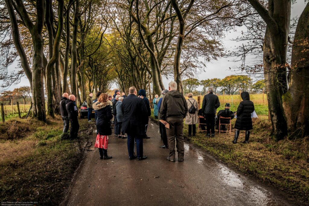 A group of people stand and talk on a tree-lined rural road with wet pavement, bordered by grass and fields on both sides, under a cloudy sky. Some people wear coats and hold umbrellas.