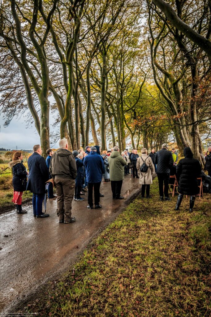 A group of people stand on a tree-lined path in autumn, many wearing coats and hats. The ground is wet, and the scene appears to be a gathering or ceremony in a rural outdoor setting.