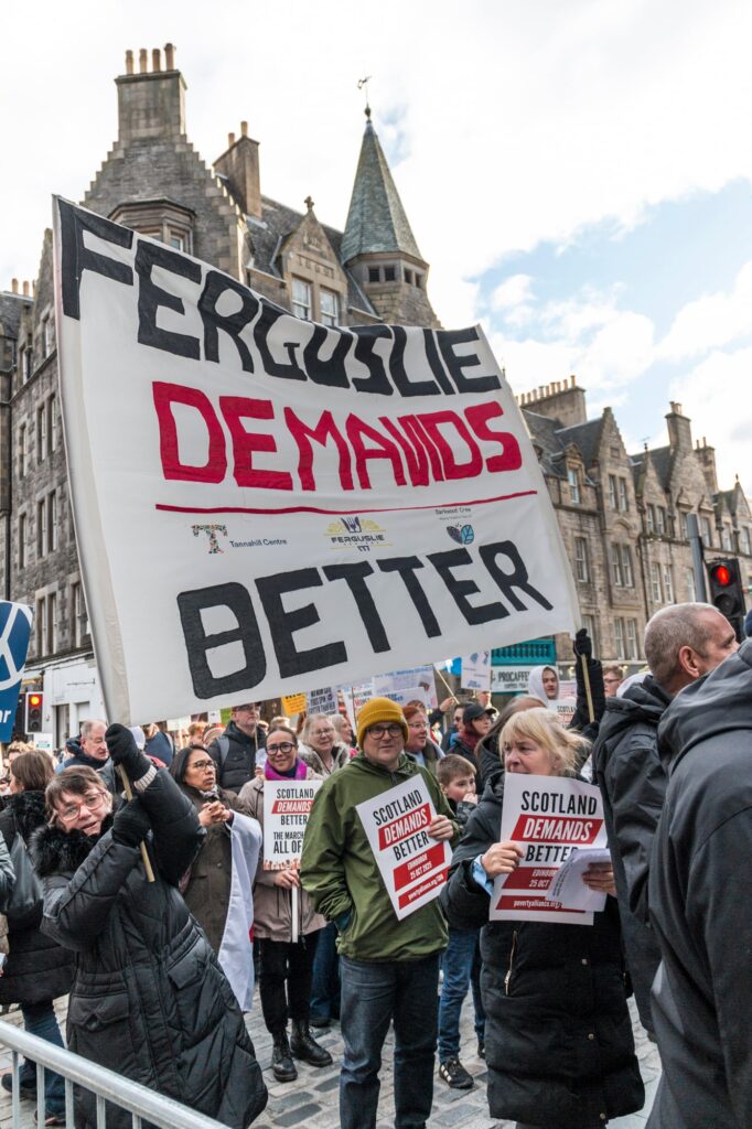 A crowd of protesters on a city street hold signs, including a large banner reading "FERGUSLIE DEMANDS BETTER," and smaller signs stating "SCOTLAND DEMANDS BETTER," with historic buildings in the background.
