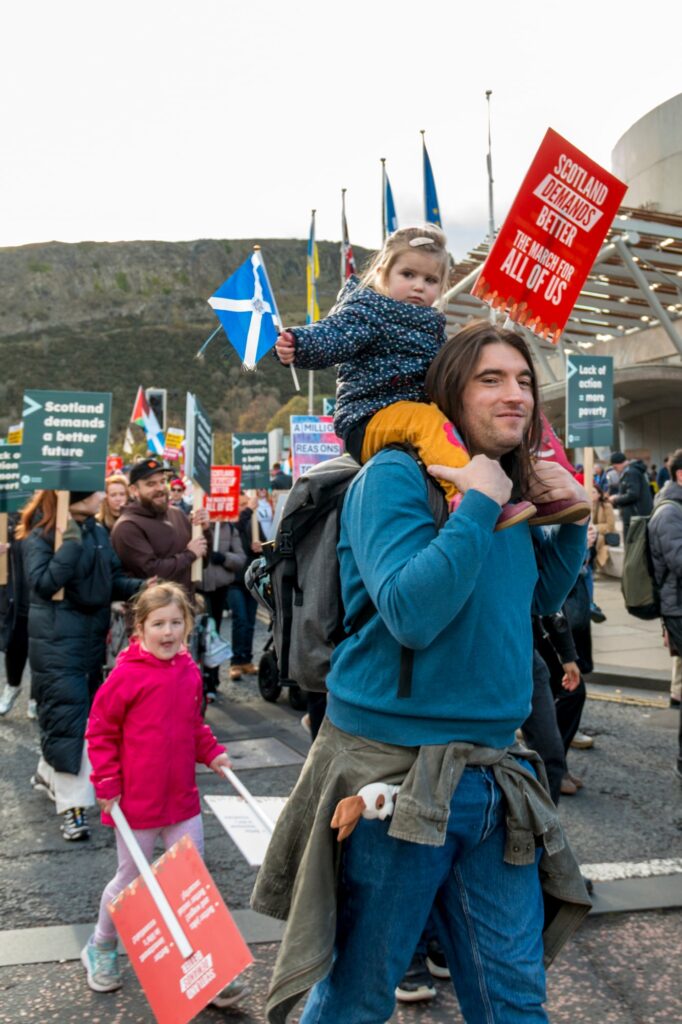 A man carries a young girl on his shoulders at a crowded protest. The girl holds a red sign that reads, "Scotland deserves better, the NHS works for all of us." Other protesters with signs and Scottish flags are in the background.