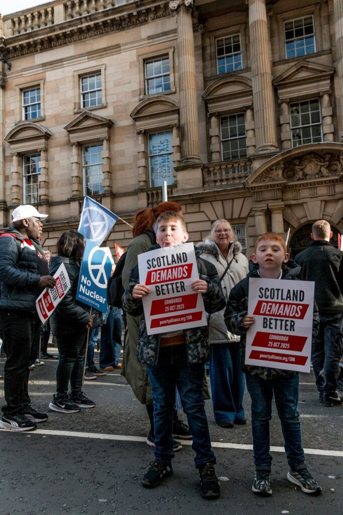 Two boys hold signs reading "Scotland Demands Better" at a protest with other people. In the background, someone holds a "No Nuclear" flag. They are standing in front of a historic building.