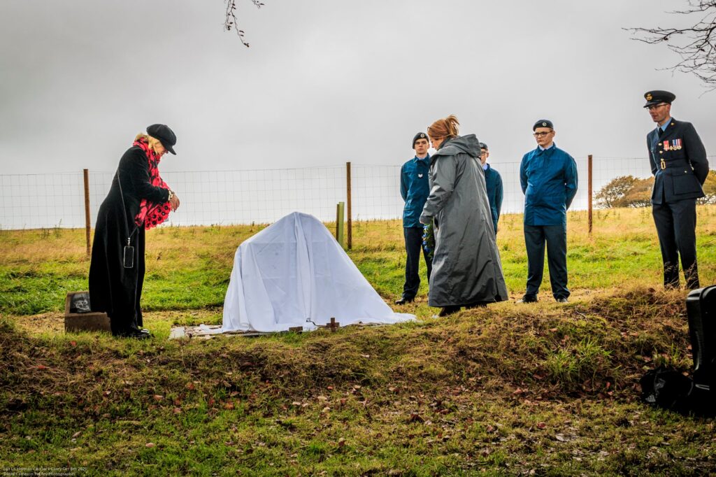 A small group stands outdoors by a grave covered with a white cloth. Three people in blue uniforms and two others in dark coats are present, with one person holding flowers. The scene looks somber and overcast.