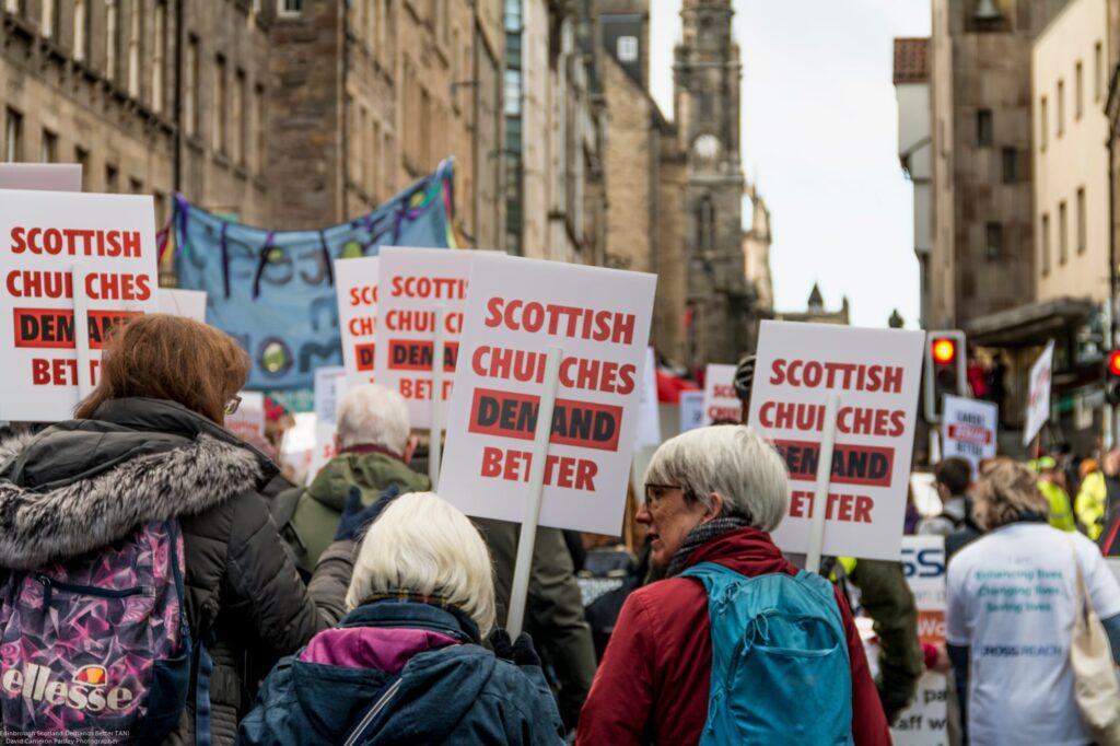 A group of protesters march down a city street holding signs that read "Scottish CHUI CHES DEN AND BET TER." The crowd is mostly older adults, and historic stone buildings line the background.