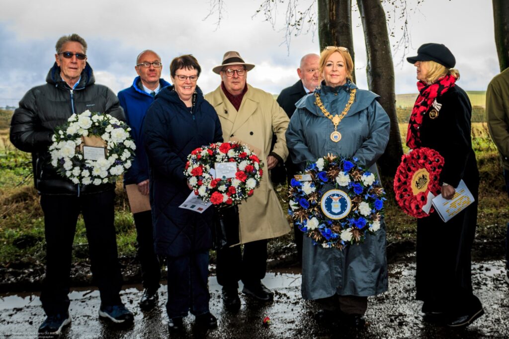 A group of people stand outdoors holding floral wreaths, some with red, white, and blue flowers. One woman wears a ceremonial chain. They appear to be participating in a commemorative or memorial event. Trees and a cloudy sky are in the background.