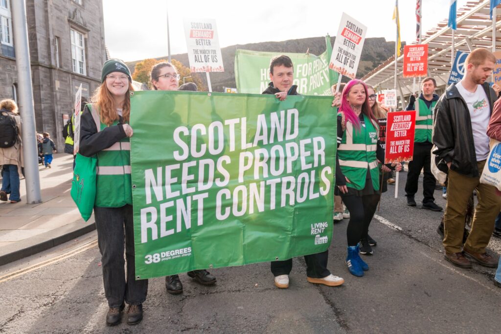 A group of people at a protest hold a large green banner that reads "Scotland needs proper rent controls." Others in the background carry signs about rent controls and tenant rights. The scene is outdoors on a city street.