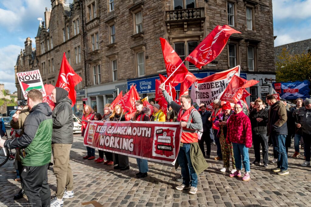 A group of protesters holding red flags and banners gather on a cobblestone street in front of stone buildings. The main banner reads "Keep Grangemouth Working" and people are dressed warmly, some wearing high-visibility vests.