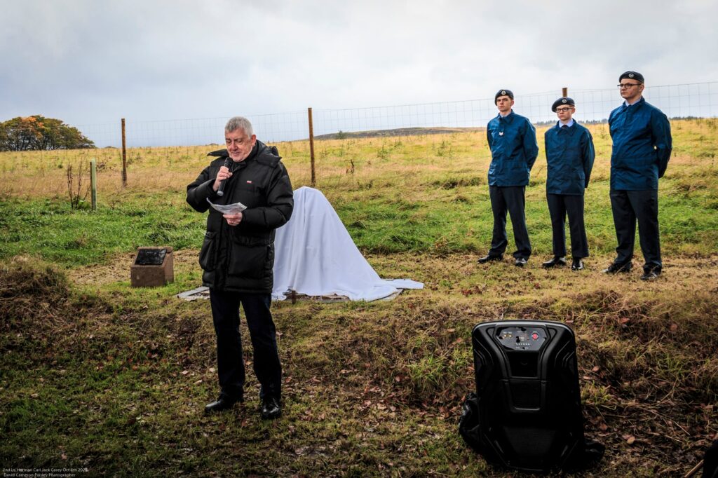 A man speaks into a microphone holding papers, standing beside a covered object outdoors. Three people in blue uniforms and berets stand behind him on grass, near a wooden fence under a cloudy sky.