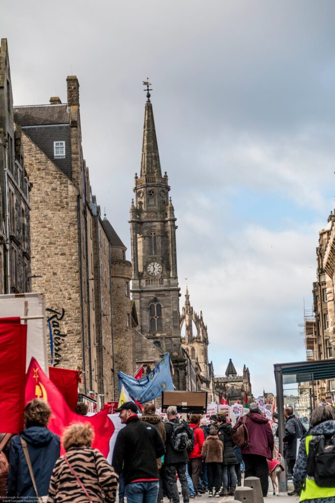A crowd of protesters holding flags and signs marches down a city street lined with old stone buildings, with a tall clock tower and cloudy sky in the background.