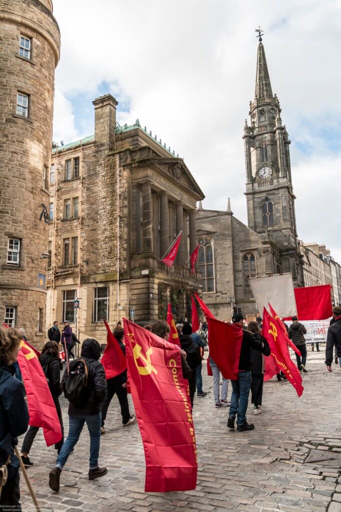 A group of people march on a cobbled street holding red communist flags. Historic stone buildings and a church tower with a clock are in the background under a cloudy sky.