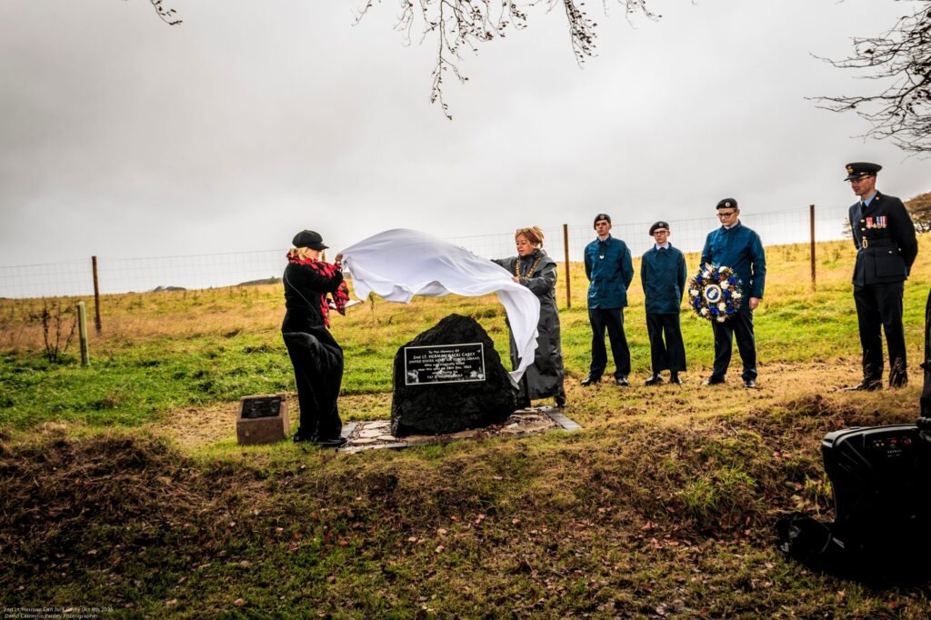 A group of people in uniform stand outdoors as two individuals unveil a memorial plaque covered with a white cloth. One holds a wreath. The setting is grassy with a wire fence and cloudy sky in the background.