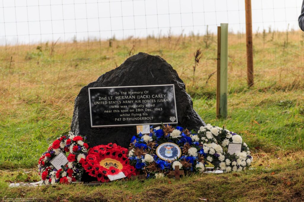 A memorial stone for 2nd Lt. Herman (Jack) Carey, decorated with wreaths and flowers, stands in a grassy field near a wire fence. The plaque commemorates his death in 1943 as a P-47 Thunderbolt pilot.