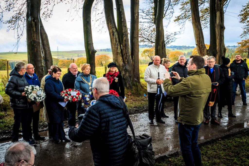 A group of people stand on a muddy path among trees, some holding wreaths of flowers. One person takes a photo while others look on. The background shows open fields under a cloudy sky.