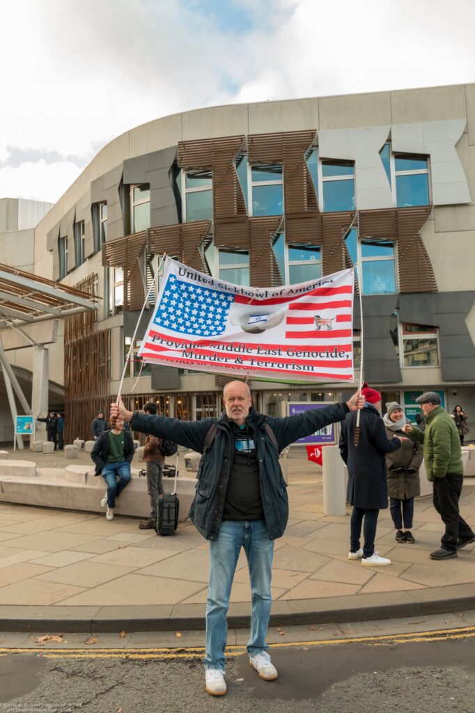 A man stands with arms outstretched holding a large American flag with altered text in front of a modern building, while several people and a suitcase are nearby on the sidewalk.