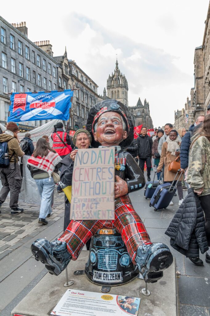 A colorful statue of a smiling man in Scottish attire sits on a busy street, holding a protest sign reading, "NO DATA CENTRES WITHOUT ENVIRONMENTAL IMPACT ASSESSMENT," with a crowd and historic buildings in the background.