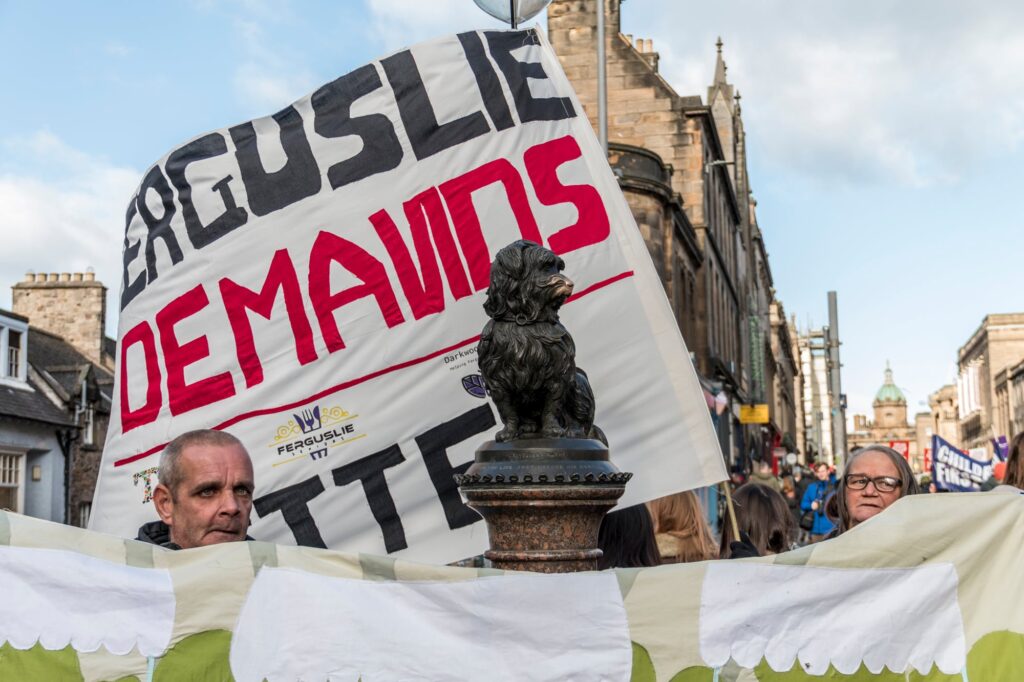 Two people hold a large protest banner with the word "DEMANDS" in red behind a small dog statue on a busy city street with old buildings in the background.