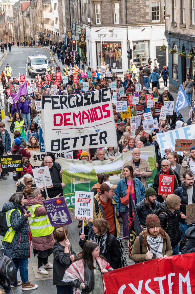 A large group of protesters march down a city street holding signs, including a prominent one that reads "Ferguslie Demands Better." The crowd appears diverse and engaged in a demonstration.