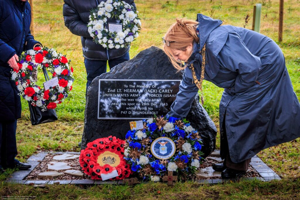 A woman in a raincoat places a wreath at a memorial stone commemorating 2nd Lt. Herman J. Carey, surrounded by wreaths and flowers on a grassy area, with others holding wreaths nearby.