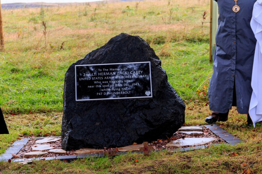 A memorial plaque on a large black stone honors 2nd Lt. Herman “Jack” Carey, a US Army Air Forces pilot, surrounded by grass and partially visible people standing nearby.