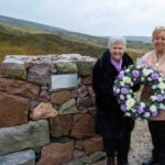 Two women stand beside a stone memorial in a grassy, rural landscape. One woman holds a wreath of white and purple flowers. The weather appears overcast and misty.